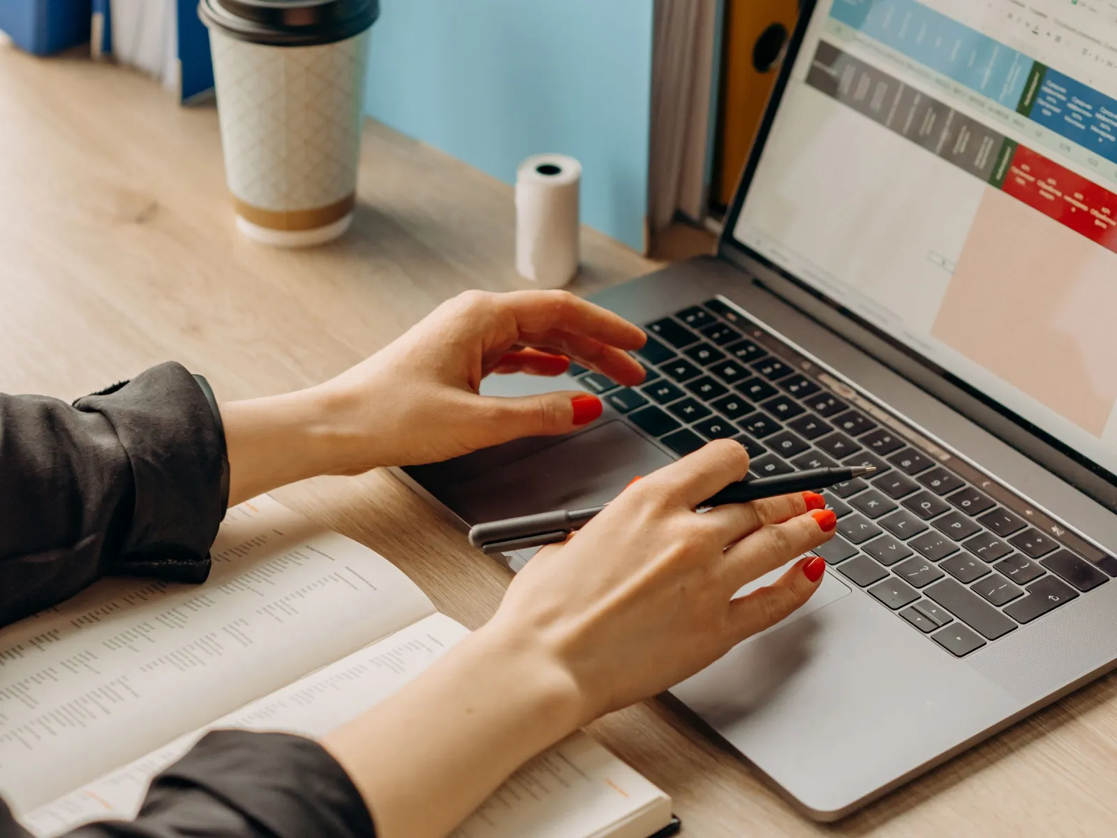 Hands at a laptop showing a spreadsheet, with ring binders and coffee nearby