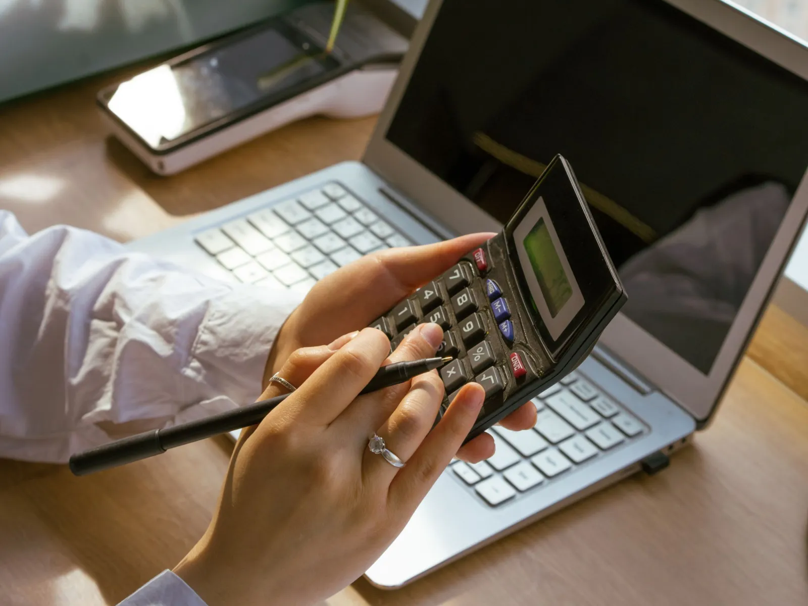 Person holding a calculator and pen in front of a laptop by a sunny window