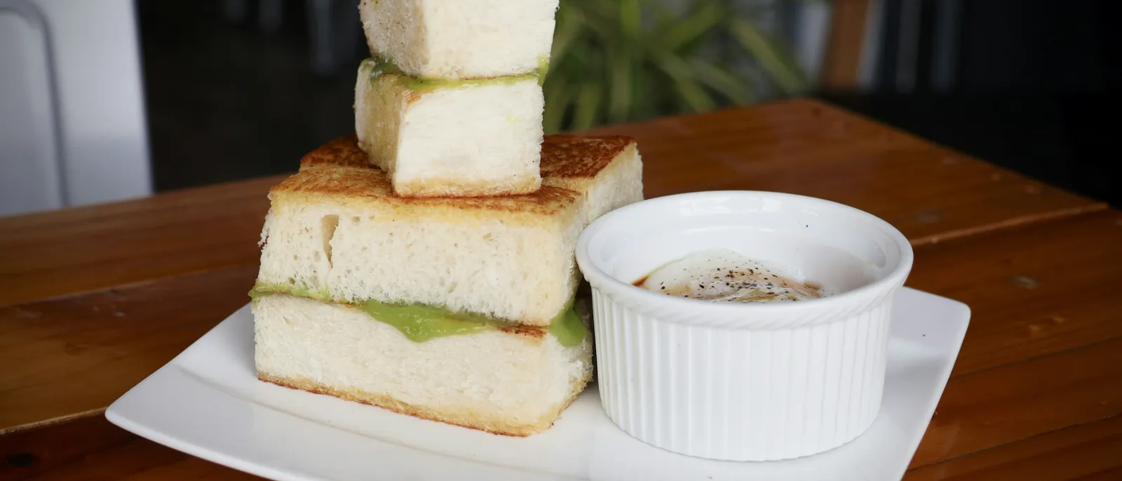 Stacked kaya toast with pandan spread and soft-boiled eggs in a ramekin on a wooden kopitiam table