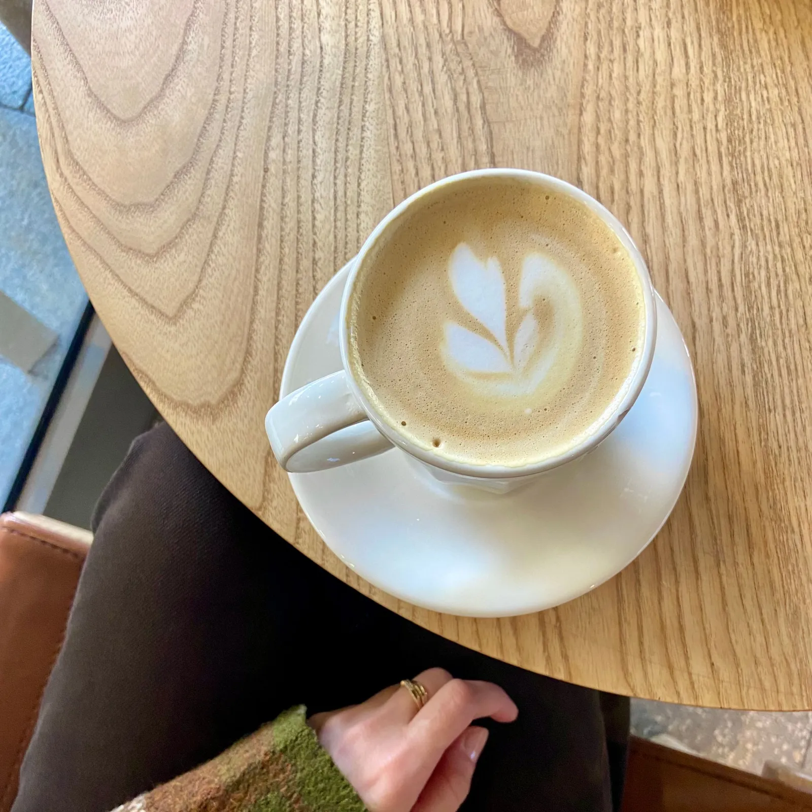 Heart latte art in a white cup on a round wooden cafe table beside a knit sweater