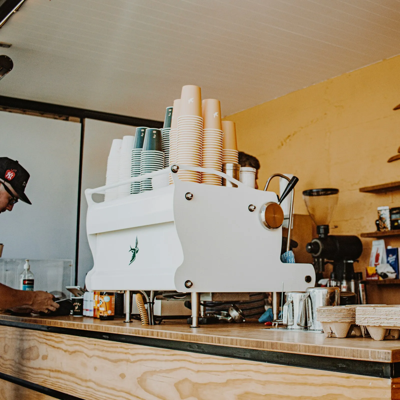 Coffee counter with white espresso machine, stacked paper cups and wooden shelves on a yellow wall