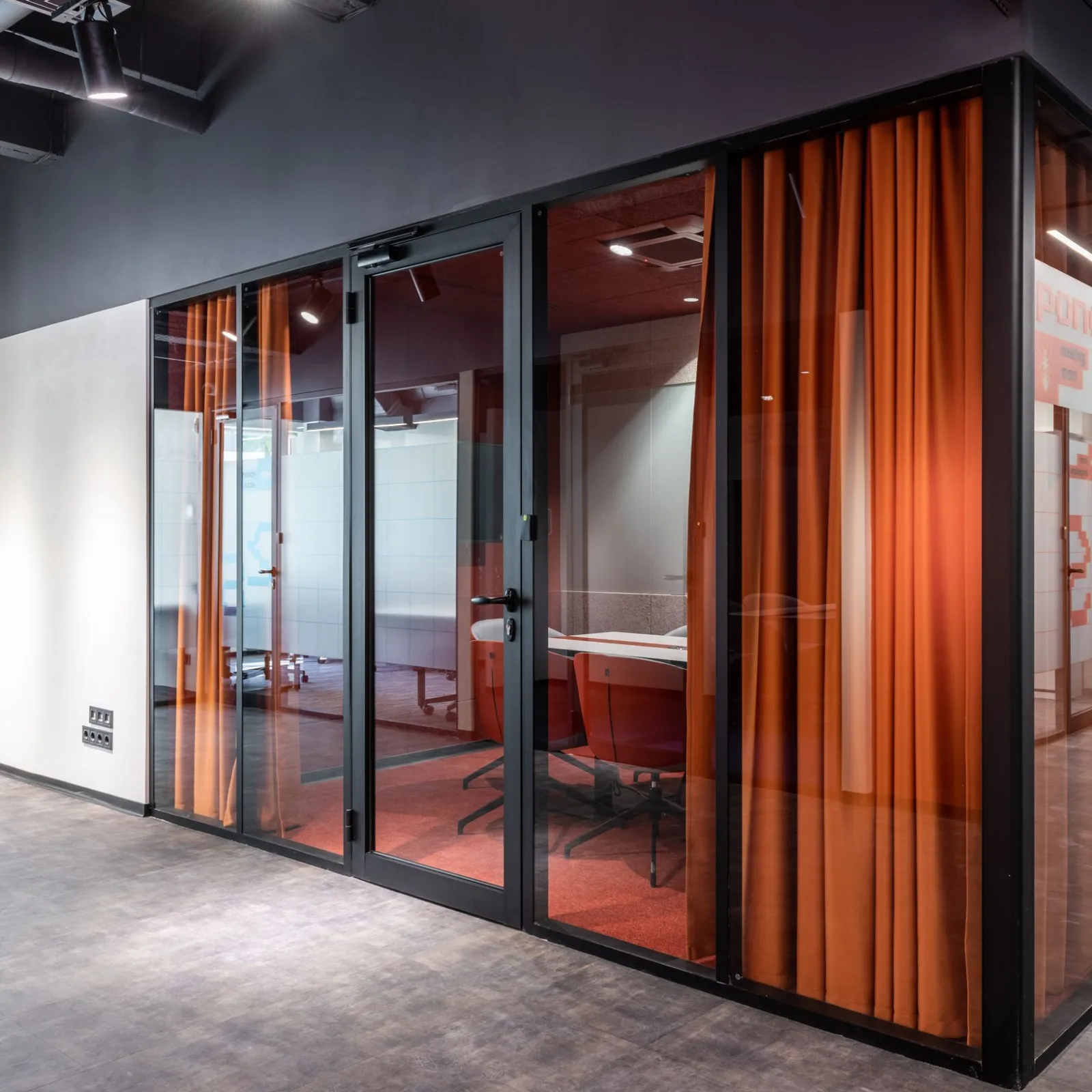 Corner meeting room with black-framed glass walls and orange curtains under a dark industrial ceiling