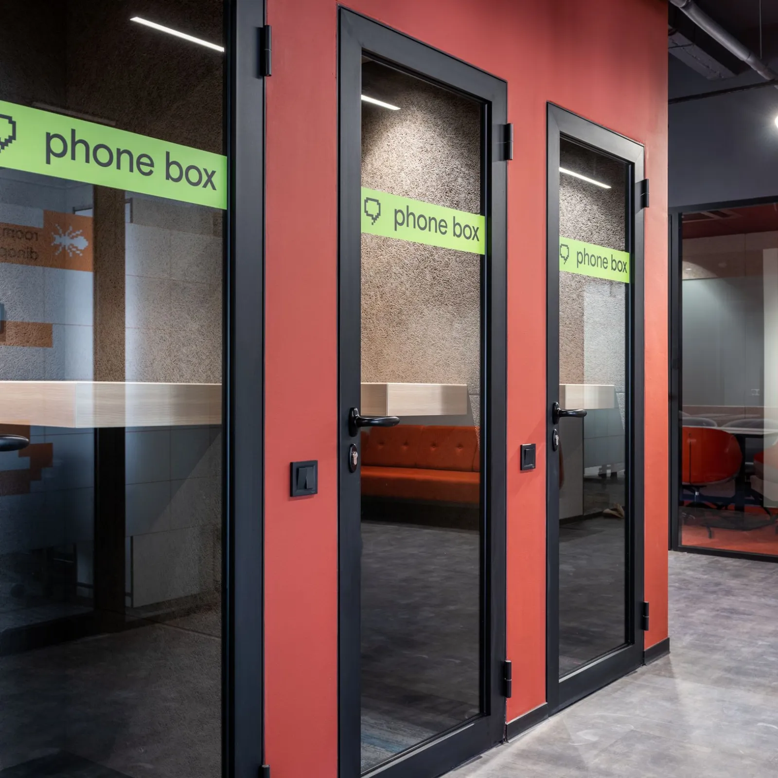 Row of glass phone booths with green phone box signs along a red and black coworking corridor