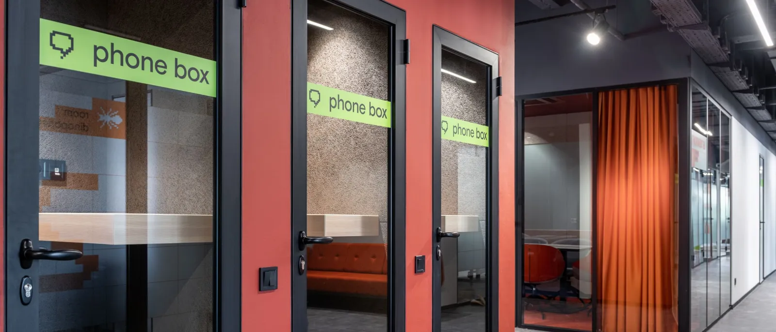 Row of glass phone booths with green phone box signs along a red and black coworking corridor