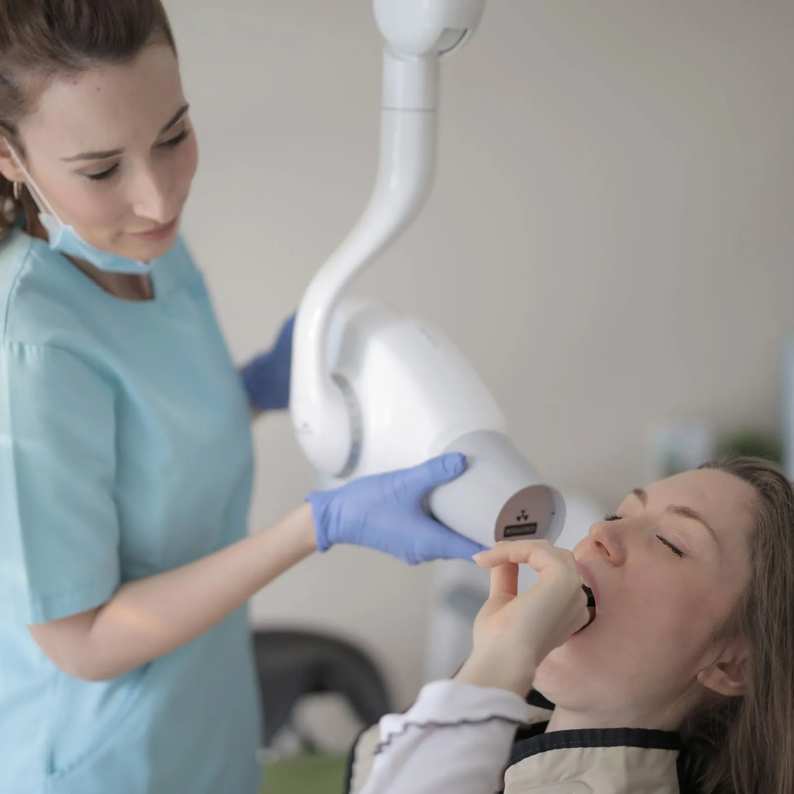 Hygienist in scrubs positioning an intraoral X-ray arm over a reclined patient