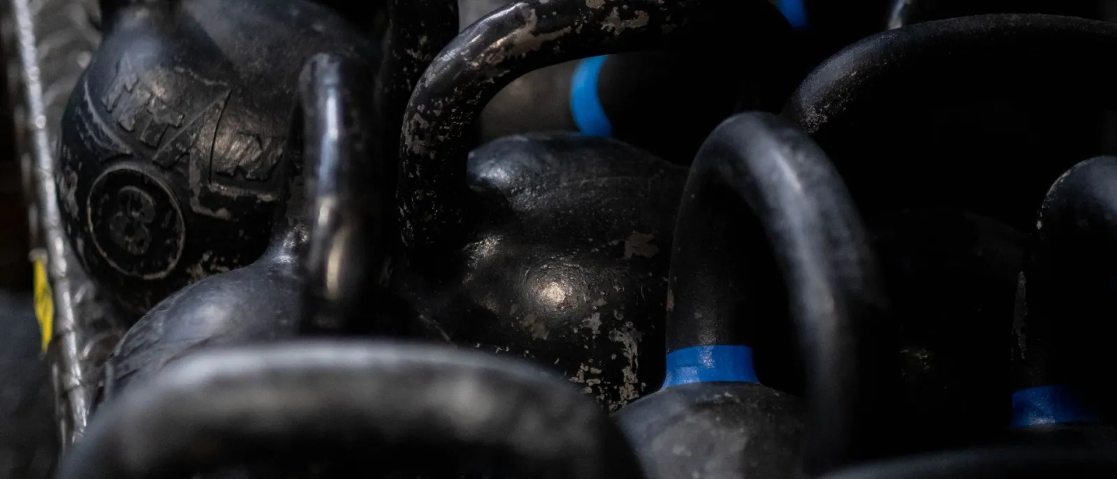 Worn black competition kettlebells crowded together on a dark gym rack