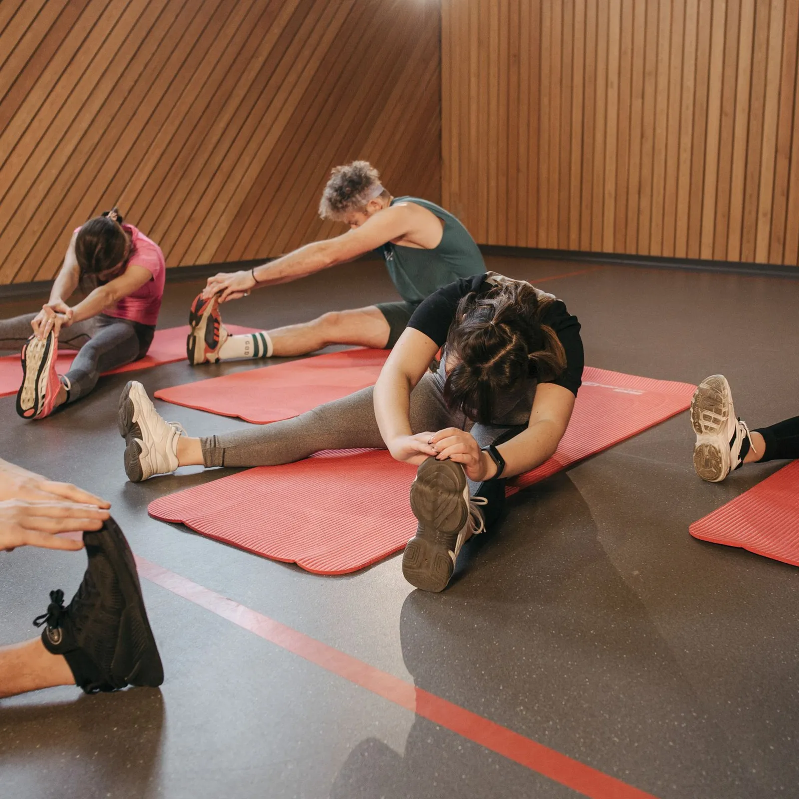 Group fitness class doing seated hamstring stretches on red mats in a wood-walled studio