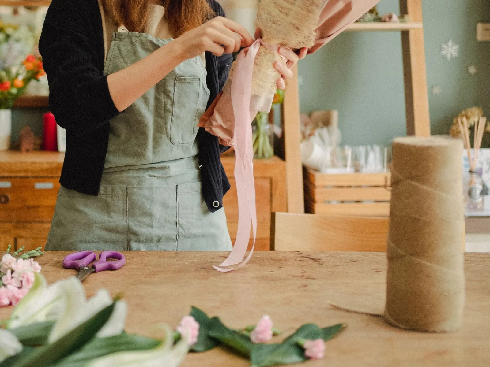 Florist in a sage apron tying pink ribbon around a paper-wrapped bouquet at a wooden workbench