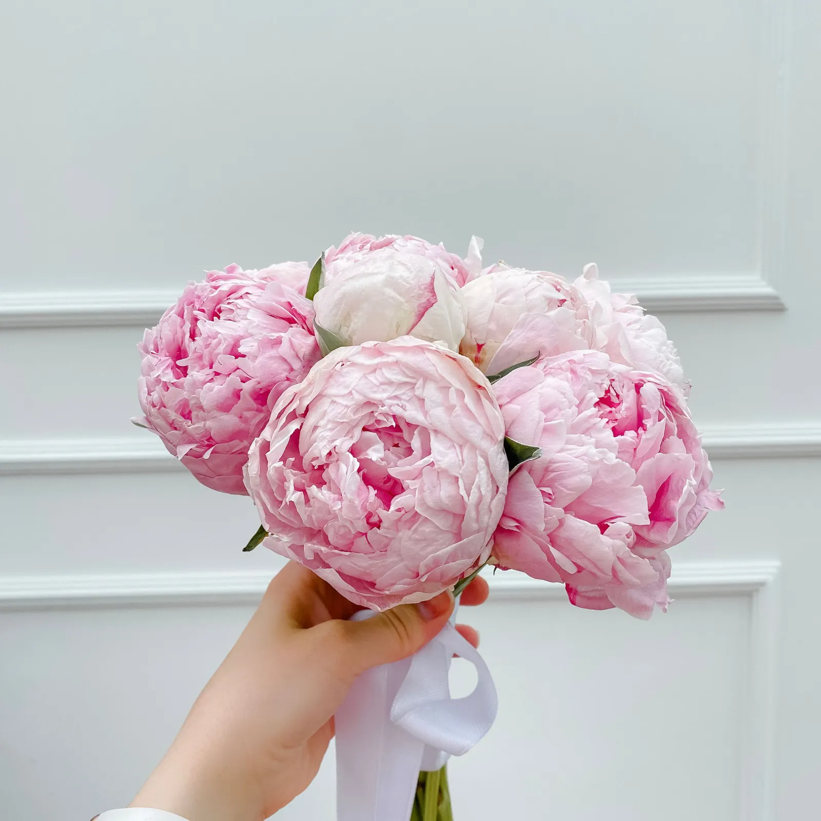 Hand raising a pink peony bouquet tied with white ribbon against a white paneled wall