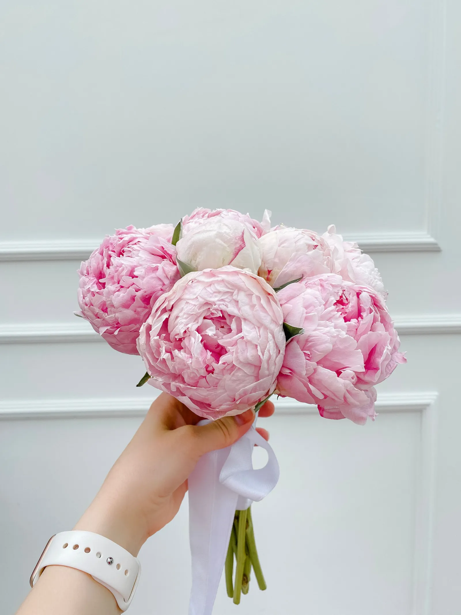 Hand raising a pink peony bouquet tied with white ribbon against a white paneled wall