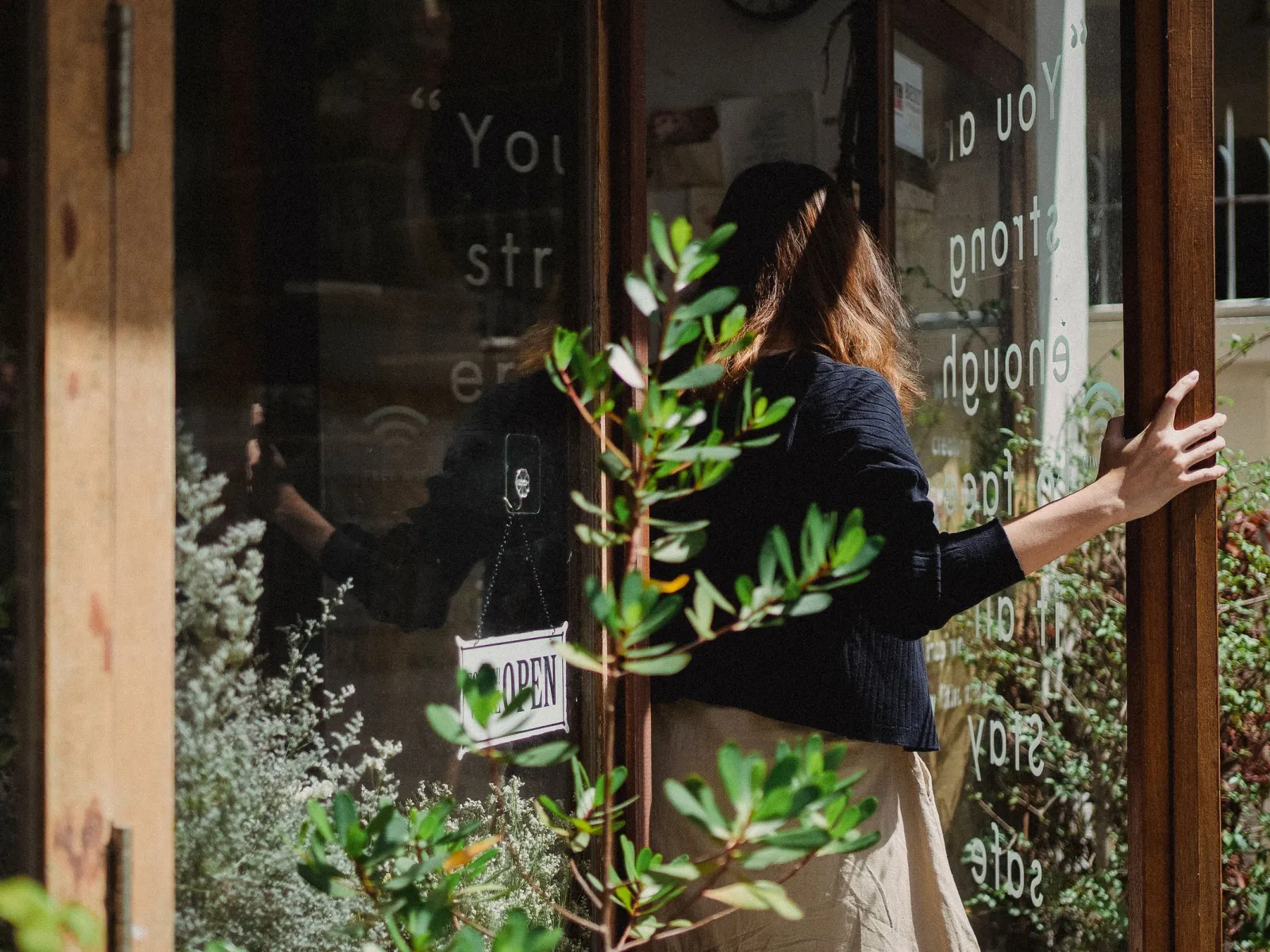 Woman seen from behind opening the glass door of a sunlit flower shop