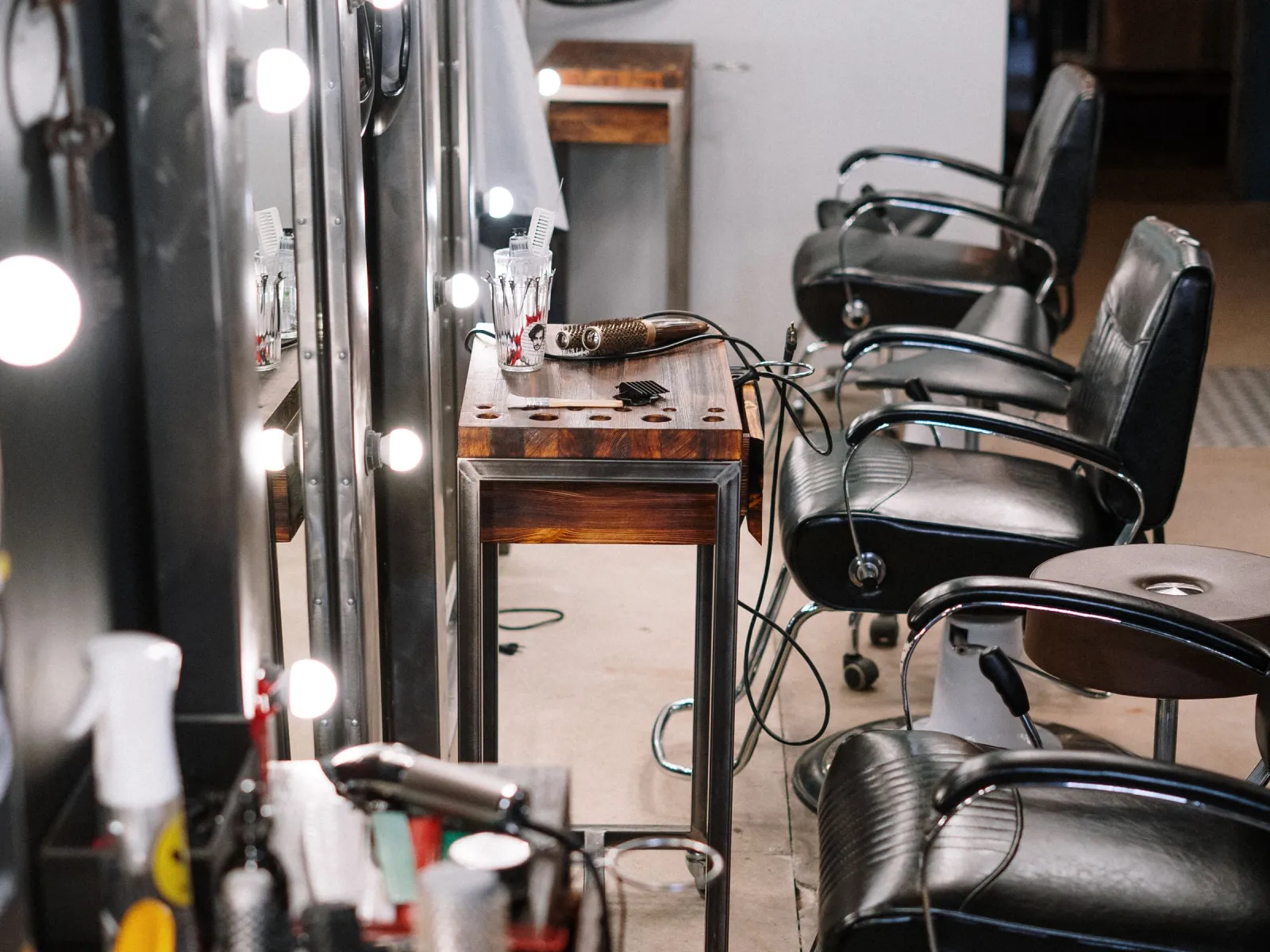Salon styling stations with bulb-lit mirrors, black leather chairs, and brushes on wooden consoles