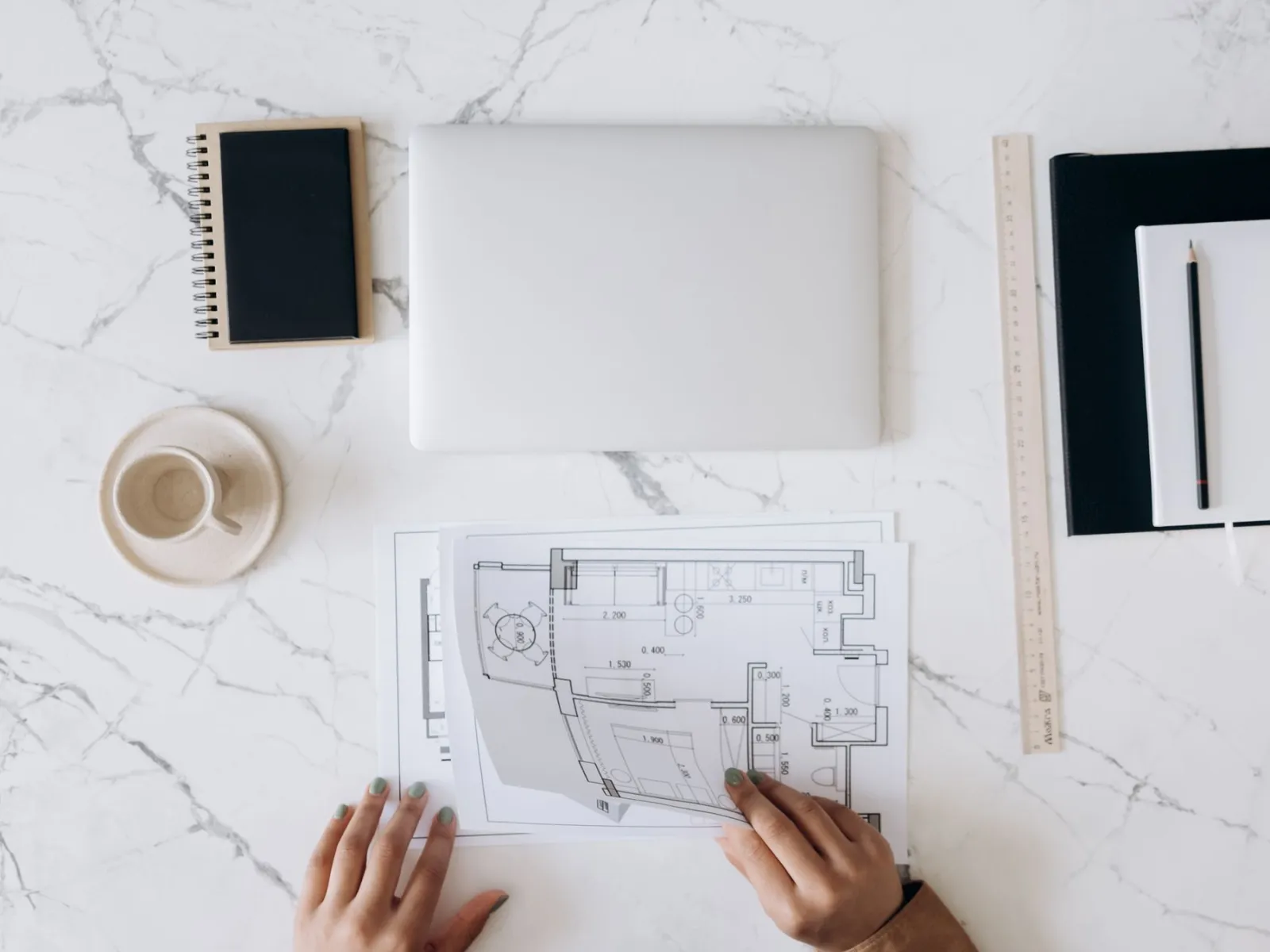 Overhead view of hands reviewing a floor plan at a marble desk with laptop, ruler and notebooks