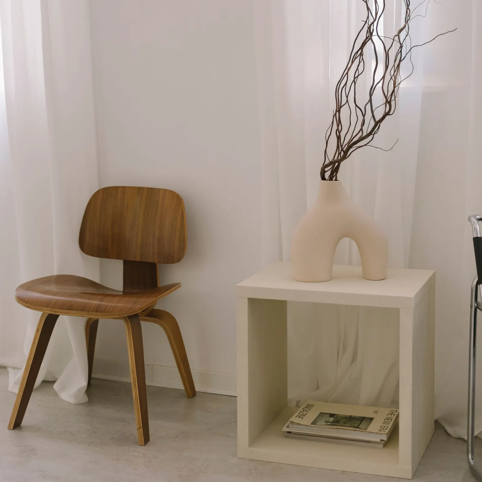 Minimalist corner with molded plywood chair and sculptural vase of branches on a cream cube table