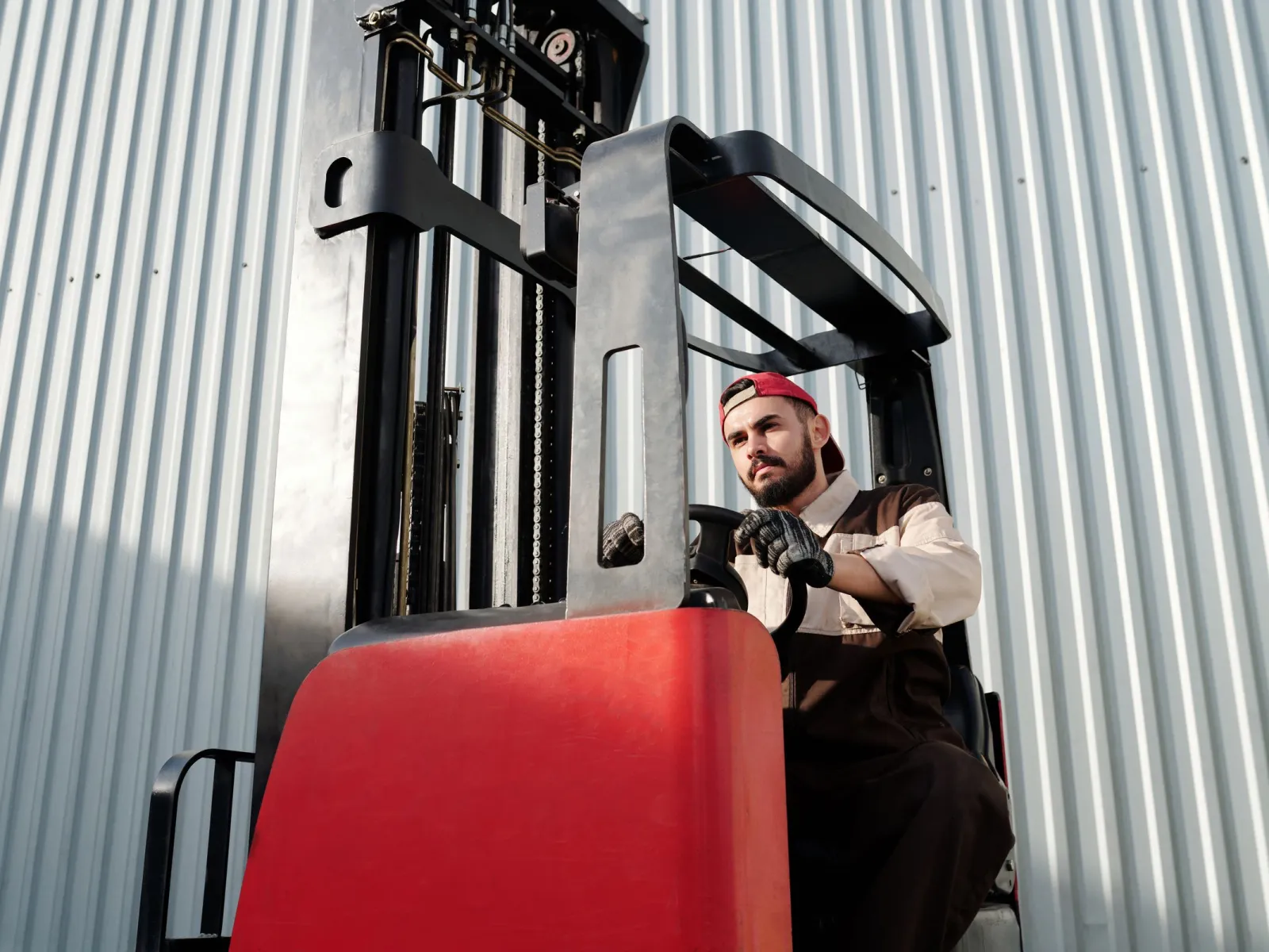 Operator in a red cap driving a red forklift beside a corrugated warehouse wall