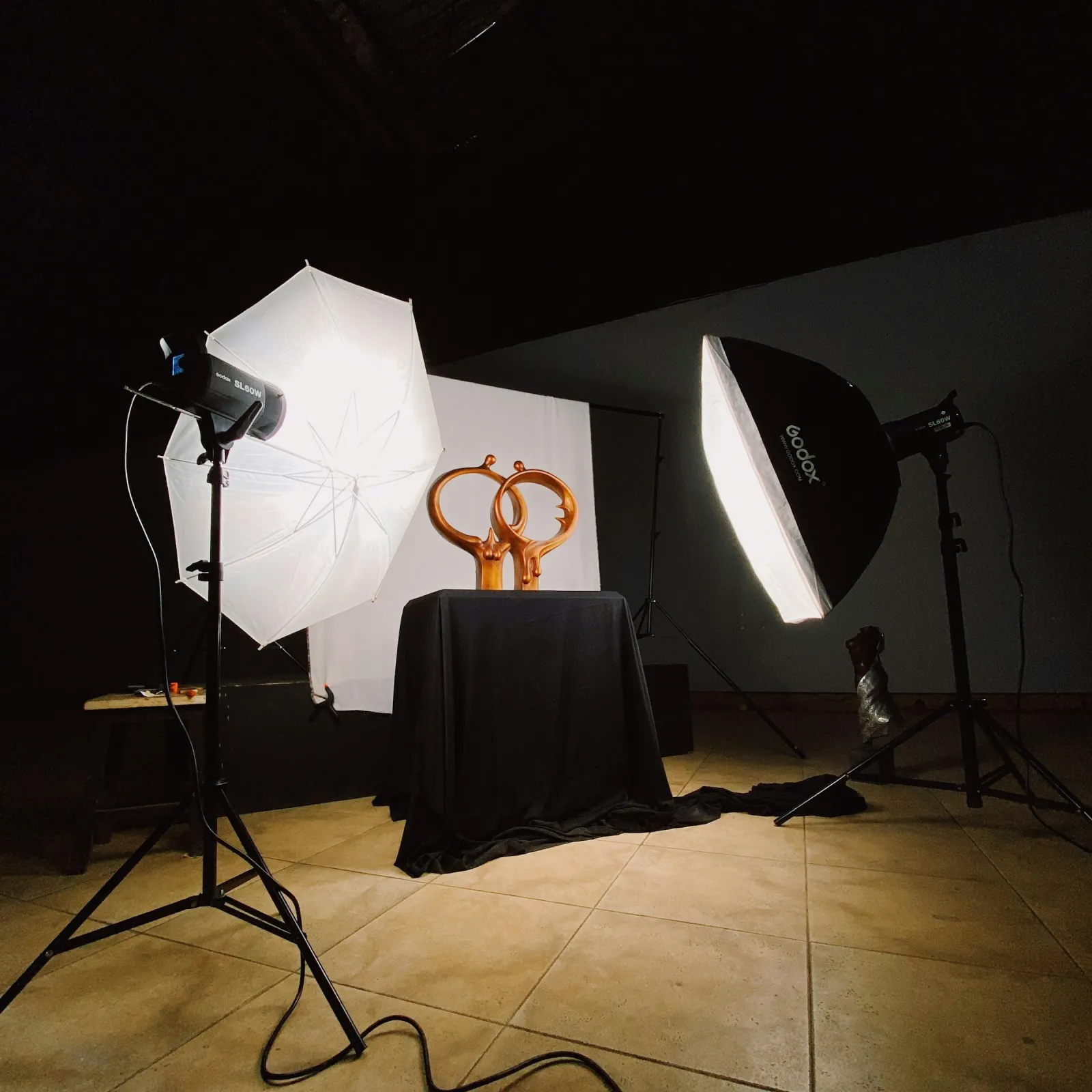 Umbrella and softbox lights aimed at bronze sculptures on a black-draped product table