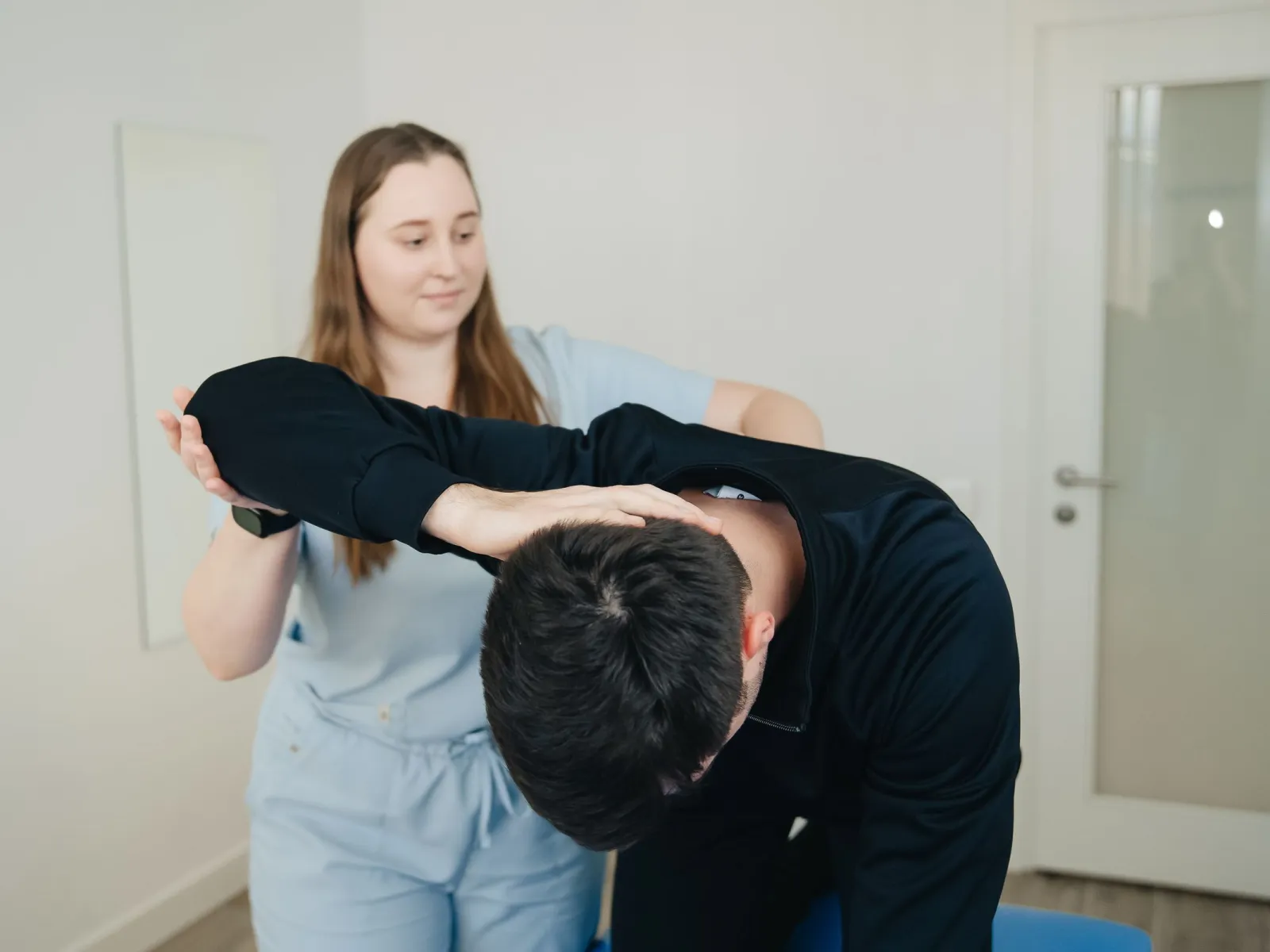 Physiotherapist guiding a man through a forward-bend mobility exercise in a bright clinic room