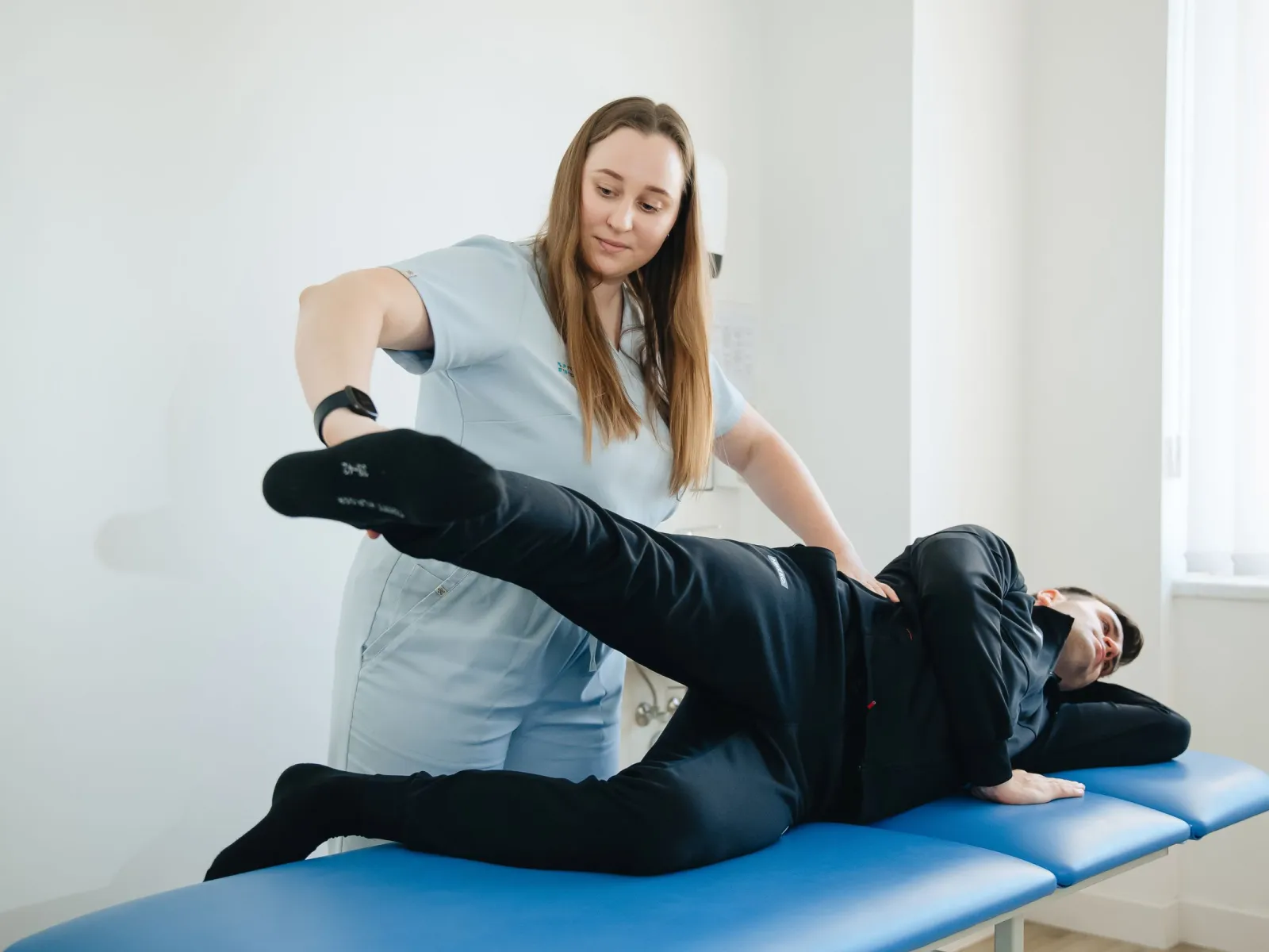 Physiotherapist supporting a patient's raised leg during side-lying therapy on a blue treatment table