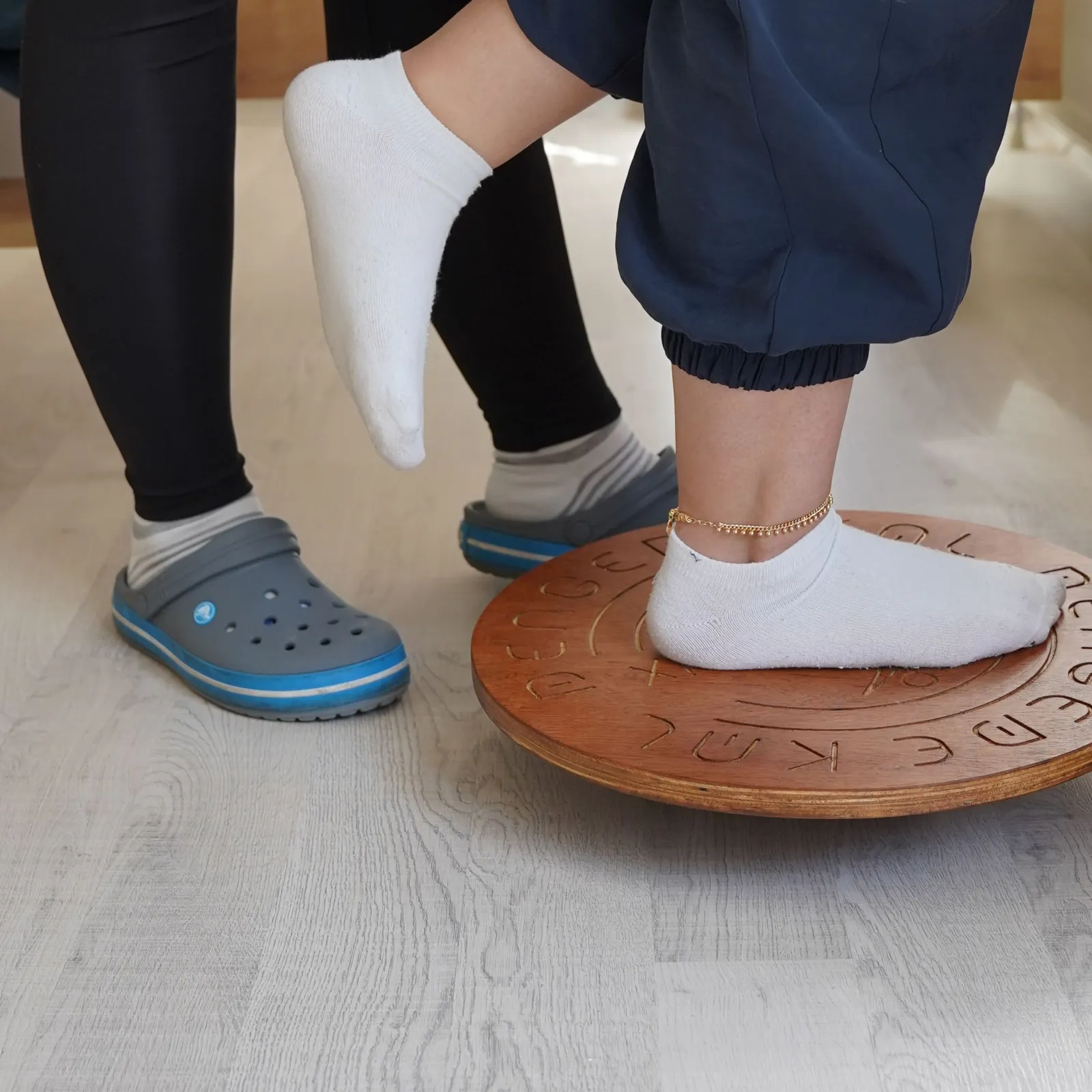 Close-up of a patient's feet training on a wooden balance board beside a physiotherapist