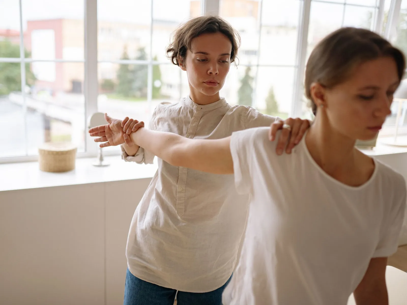 Therapist guiding a woman's outstretched arm during a posture exercise by large studio windows