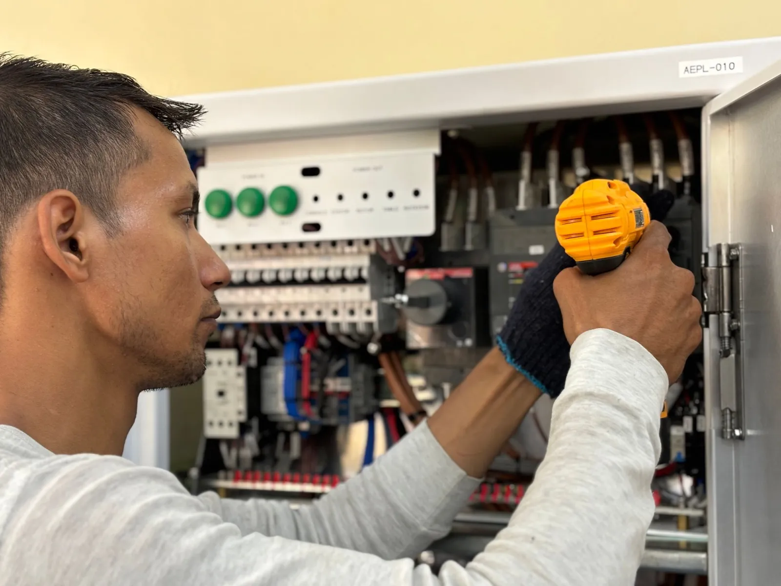 Electrician steadying a cordless driver inside an open control panel with green indicator buttons