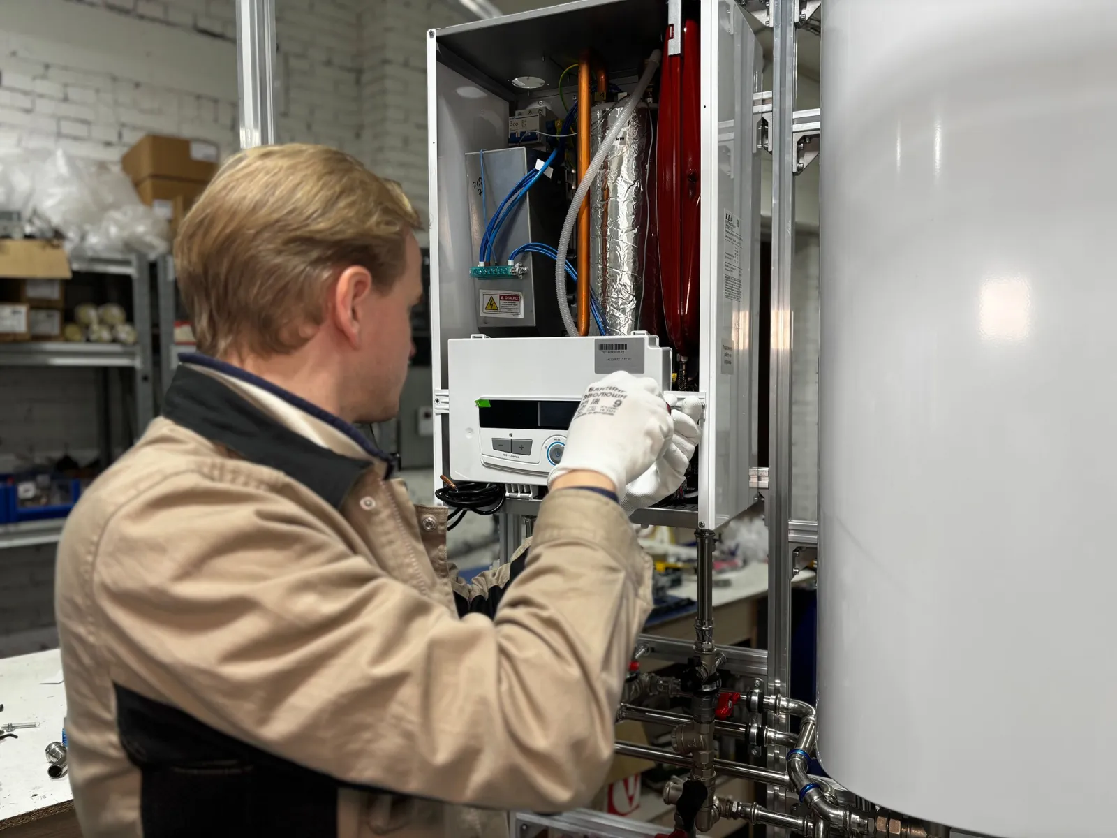 Technician in gloves fitting the control module of a wall-hung boiler beside a storage tank