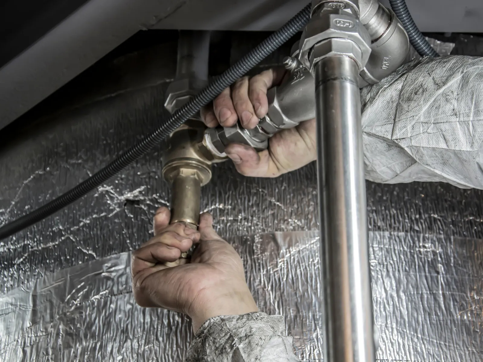 Plumber's hands tightening steel pipe fittings beneath equipment lined with foil insulation