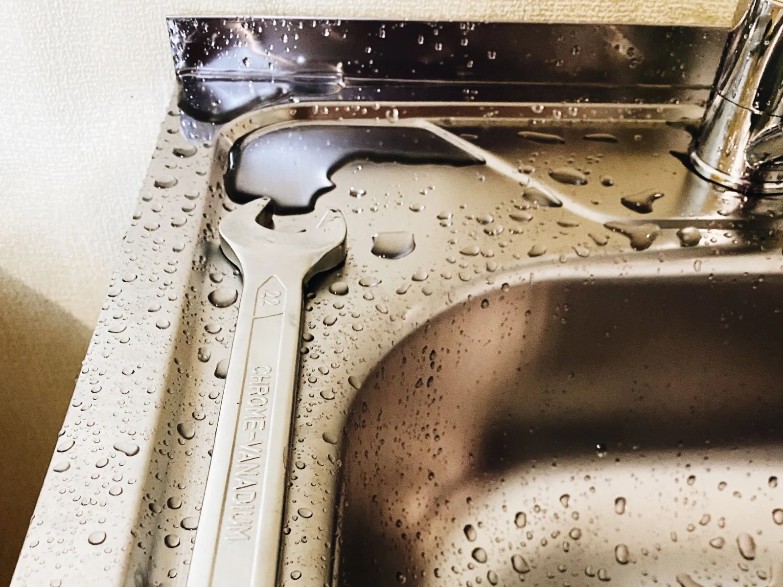 Chrome spanner lying on a wet stainless steel sink beside the base of a kitchen tap